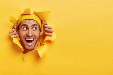 Satisfied cheerful guy with broad toothy smile, sticks head through ripped hole in yellow paper...