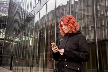 Beautiful, young woman is looking into the phone, against the gray modern building background. Gloomy autumn city in November.