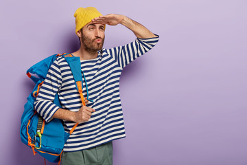 Serious backpacker keeps lips folded, looks far away with hand on forehead, carries blue rucksack on shoulder, isolated over purple background with blank space aside. Tourism and discovering concept