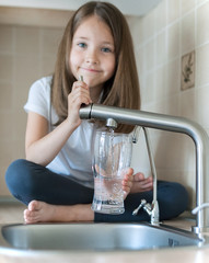Portrait of a little caucasian girl gaining a glass of tap clean water. Kitchen faucet. Cute curly...
