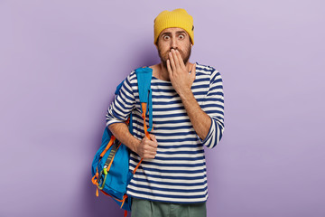 Speechless emotive backpacker holds palm on mouth, cannot believe eyes, has active lifestyle,...