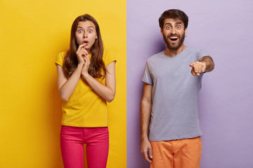 Photo of shocked couple gaze straightly at camera with bated breath, happy unshaven man in casual wear points at something, stands over purple background, impressed woman looks with excitement