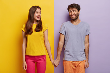 Romantic couple in love have date, hold hands, look positively at each other, feel support, walk together. Positive man poses over purple background, woman on yellow. Contrast. Relationship concept