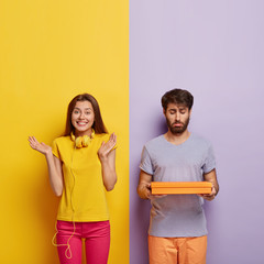 Happy smiling young woman clasps hands, glad to receive present from boyfriend, pose together against colorful background. Puzzled unshaven attractive man carries small box, has sad facial expression