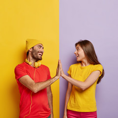 Glad boyfriend and girlfriend touch by their palms, smile positively, agree about something, dressed in red and yellow casual t shirts, enjoy free time together, stand against colored background