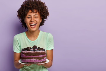 Happy birthday girl laughs joyfully, holds big tasty fruit cake, likes eating sweet food, improves...