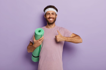 Glad unshaven man makes okay gesture, keeps thumb up, holds rolled up karemat, shows he liked yoga...