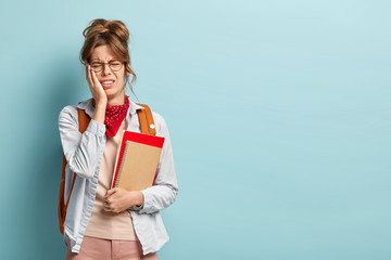 Photo of dissatisfied stressful tired schoolgirl carries spiral notepad and book, wears round spectacles, bandana, shirt, stands against blue wall with blank copy space. Negative emotions concept