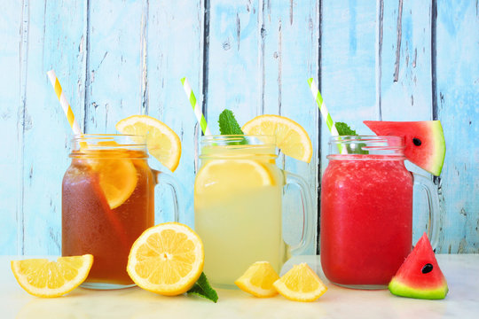 Variety of summer drinks in mason jar glasses with fruit against a blue wood background. Iced tea, lemonade and watermelon juice.