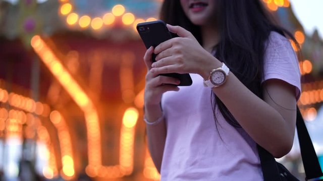 Asian beautiful woman using smartphone doing chatting with smartphone at amusement park carnival near carousel. Woman happy travel night funfair festival Slow motion.