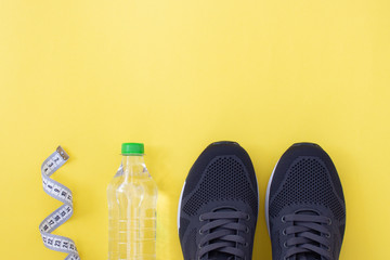 Fitness flat lay. Healthy lifestyle and sport concept. Black sneakers, tape measure and bottle of water on a yellow background.