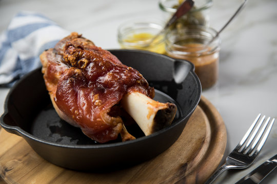Gegrillte Schweinefleisch Haxe in gusseiserne Pfanne auf Holzbrett mit Cornichons, süßen und mittelscharfen Senf auf Marmor Hintergrund