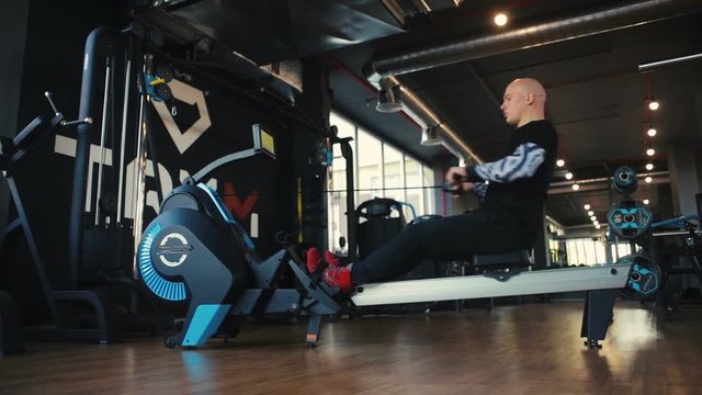 handsome bald man in black sportswear uses rowing machine to strengthen arm and back muscles close view