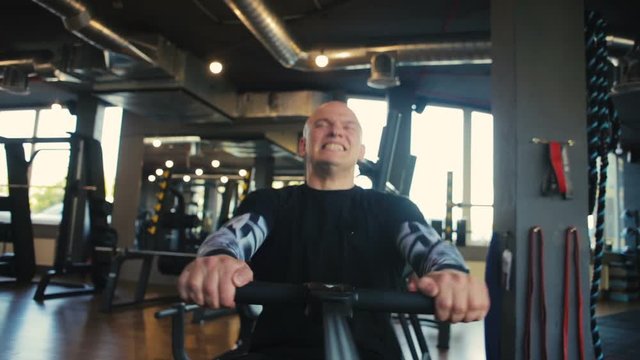strong bald man trains hard on rowing machine and has cardio workout in empty modern gym extreme close view