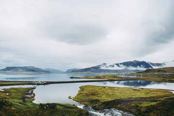 Obraz premium Colorful Icelandic landscapes under gray heavy sky. Green wet grass, beautiful lake, and snowy tops of the mountais reflected in the lake water. Kirkjufellsfoss, Snaefellsnes peninsula.
