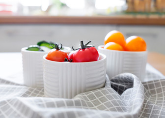 Red and yellow cherry tomatoes and sliced cucumber in white bowls on the kitchen countertop. 