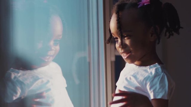 Curious afro-american little girl touching the window looking at lovely sunset. Portrait of amazing kid having fun at home.