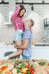 Happy young couple have fun in modern kitchen while preparing vegetables. Man holding his wife on hands.