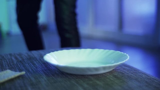 Somebody dressed in black pants aggressively hits several times white plate with two small buns and few thin slices of bread with thick pink stick, in light room.