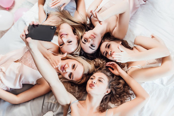 Photo of five, cheerful girls in night robes lying on bed, making selfie, looking  and posing at camera.. Hen party.