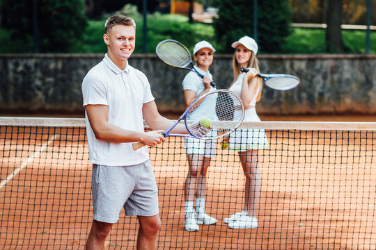Profile picture of young happy man in sportswear, playing tennis, waiting for the serve , with two girls behind. On the court.