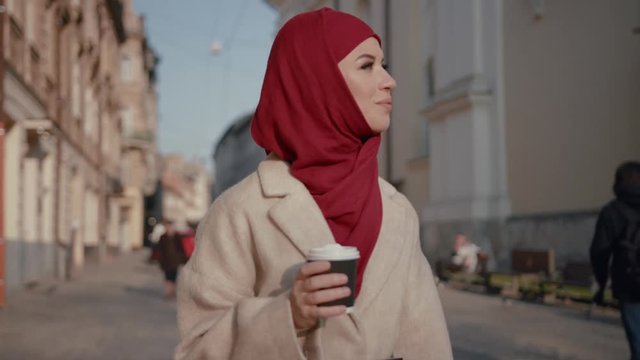 Happy romantic arab woman holding cup drinking coffee walking on the beautiful street in the city. Dream vacation, coffee break.