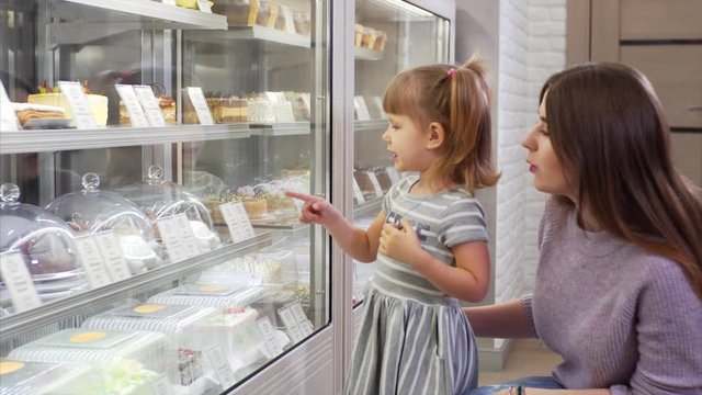 Young mother with cute daughter choose something tasty for dessert near glass shop window in coffee shop. 
