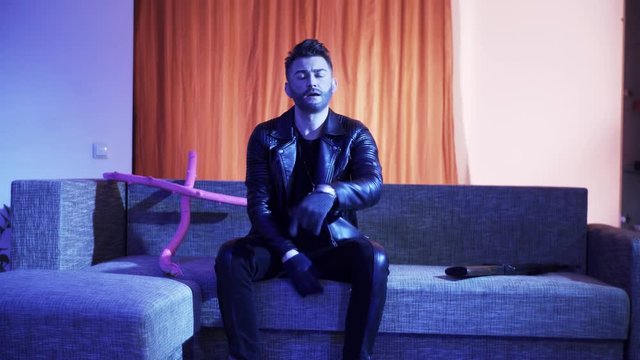 Handsome bearded man with stylish haircut, wearing black leather jacket, gold cross with chain and gloves, sits on modern grey sofa with wooden pink cross and machine gun, sings and looks at camera.
