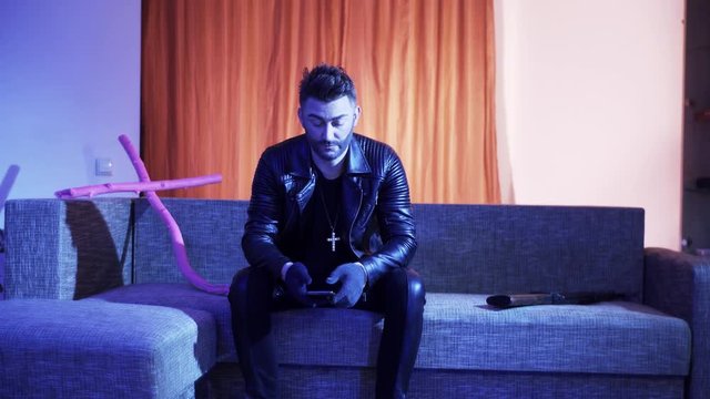 Attractive bearded man with stylish haircut, wearing black leather jacket, gold cross with chain and gloves, sits on modern grey sofa with wooden pink cross and machine gun, sings and looks at camera.