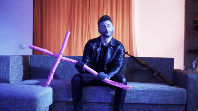 Cute bearded man with stylish haircut, wearing black leather jacket, gold cross with chain and gloves, sits on modern grey sofa with wooden pink cross and machine gun, sings and looks at camera.