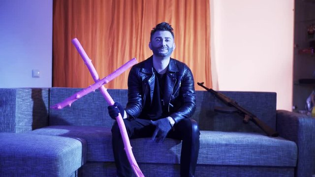 Nice-looking bearded man with cute haircut, wearing black leather jacket, gold cross with chain and gloves, sits on modern grey sofa with wooden pink cross and machine gun, sings and looks at camera.