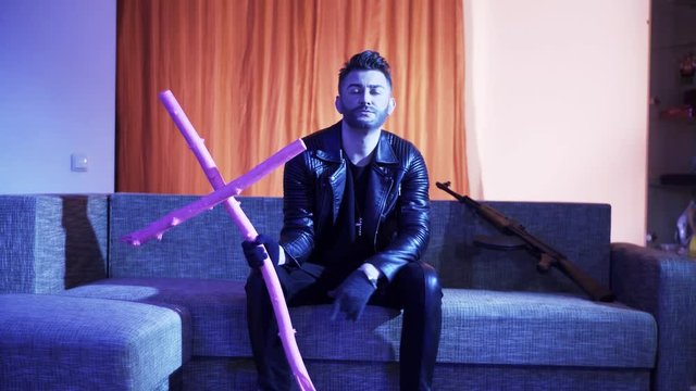 Handsome bearded man with stylish haircut, dressed in black leather jacket, gold cross with chain and gloves, sits on modern grey sofa with wooden pink cross and machine gun, sings and dances.