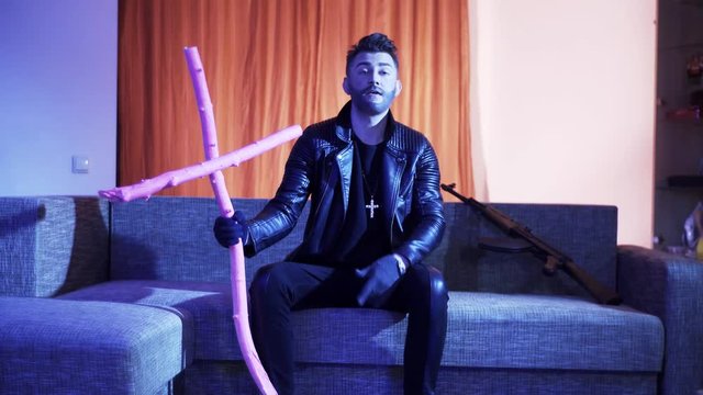 Handsome bearded male with stylish haircut, dressed in black leather jacket, gold cross with chain and gloves, sits on modern grey sofa with wooden pink cross and machine gun, sings and dances.