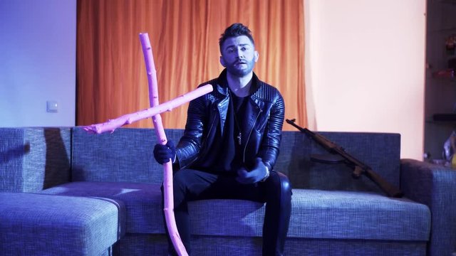 Handsome bearded guy with stylish haircut, wearing black leather jacket, gold cross with chain and gloves, sits on modern grey sofa with wooden pink cross and machine gun, sings and looks at camera.