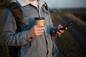 Close-up of hands of young modern guy with backpack, holding the coffee cup and using smartphone with earphones