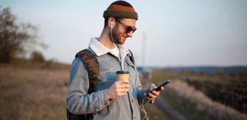 Tuinposter Portrait of young guy holding cup of hot coffee and smartphone with earphones outdoor © Lalandrew