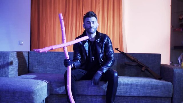 Cute bearded guy with stylish haircut, wearing black leather jacket, gold cross with chain and gloves, sits on modern grey sofa with wooden pink cross and machine gun, sings and looks at camera.