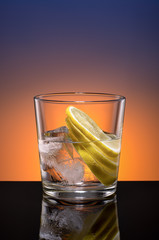 Transparent glass with water and natural ice cubes and lemon slices on orange-purple background