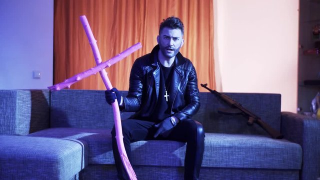 Young cute bearded man with stylish haircut, wearing black leather jacket, gold cross with chain and gloves, sits on modern grey sofa with wooden pink cross and machine gun, sings and looks at camera.
