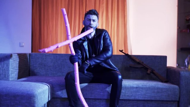 Attractive young bearded male with stylish haircut, wearing black leather jacket, gold cross with chain and gloves, sits on grey sofa with machine gun, holds wooden pink cross , dances and sings.
