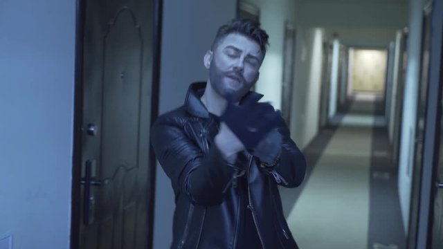 Cute bearded young man with stylish haircut, dressed in black tight leather jacket and wearing gold cross with chain, actively dances and sings looking at camera standing in long narrow corridor.