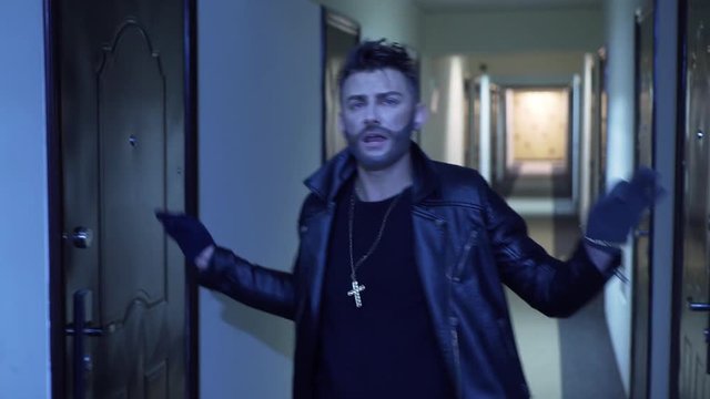 Young handsome man with stylish haircut and fake beard, dressed in black tight leather jacket and wearing gold cross with chain, actively dances and sings, standing in long narrow corridor.