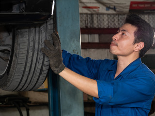 Handsome asian mechanic inspecting wheel and suspension detail of lifted automobile at repair garage.