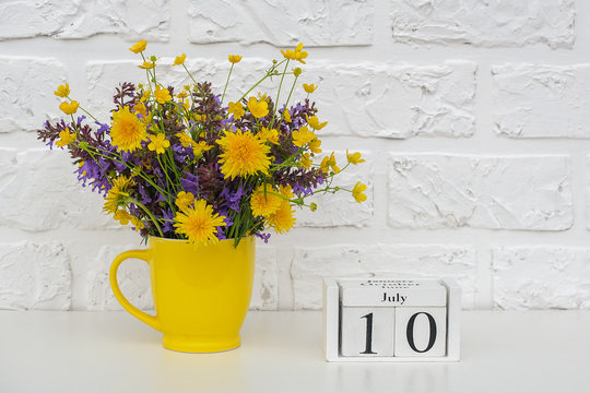 Wooden cubes calendar July 10 and yellow cup with bright colored flowers against white brick wall. Template calendar date for your design Copy space - Powered by Adobe