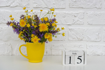 Wooden cubes calendar July 15 and yellow cup with bright colored flowers against white brick wall. Template calendar date for your design Copy space