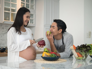 Young asian expecting pregnant couple cooking together in the kitchen at home. Pregnant woman preparing food with her husband.