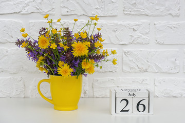 Wooden cubes calendar July 26 and yellow cup with bright colored flowers against white brick wall. Template calendar date for your design Copy space.