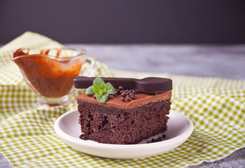 A piece of homemade chocolate cake on the plate with icing, mint leaf and chocolate spoon on the gray table