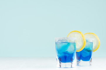 Exotic cold shot glass cocktail with yellow lemon slice, blue curacao, ice cubes, salt rim on soft light mint color background and white wood board.