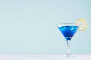 Refreshing alcohol drink blue Hawaii in misted rich wine glass with ice cubes, lemon slice in modern pastel mint beach bar interior.